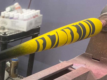 A Razorbill wood bat in the process of being painted on a lathe, with a vibrant yellow finish and large Razorbill lettering prominently displayed on the barrel.